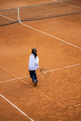 A little girl plays tennis on a red clay court. She runs and trains to learn the beautiful sport of tennis. Competitiveness, athlete, sporty child.