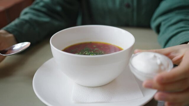 Woman gently adds a dollop of sour cream to a bowl of vibrant borscht, garnished with fresh herbs, in a well-lit setting perfect for culinary themes. Healthy Organic Borscht Soup Being Seasoned with