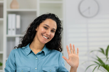 Smiling woman in a casual shirt waving hello during a video call, providing an engaging webcam view...