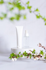 White unbranded cosmetic tube and glass bottle with dropper on marble cosmetic podium decorated with blooming apricot branches, cosmetic branding, vertical image.