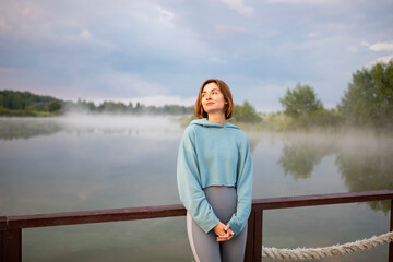 Portrait of young and attractive girl standing at the coast of the foggy river	