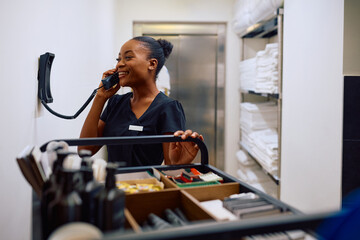 Happy black housekeeper talking on the phone in hotel laundry room.