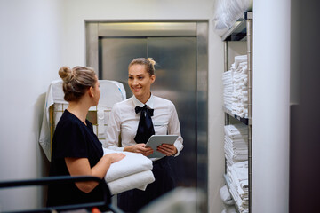 Happy housekeeping supervisor and chambermaid communicating in hotel laundry room.