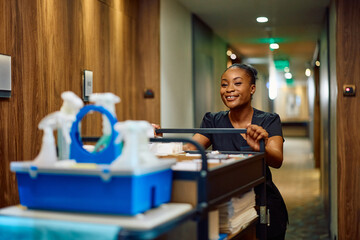 Happy black housekeeper with chambermaid's trolley in hotel hallway.