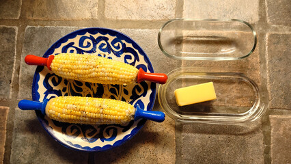 hot buttered sweet corn on the cob with sea salt and black pepper. Clear glass butter dish with butter also pictured.  - 776327465