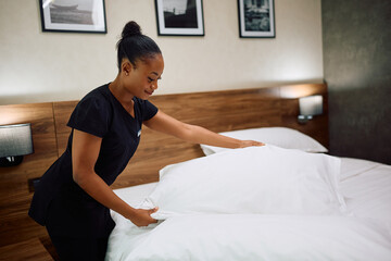 Smiling black maid changing bedding in hotel room.