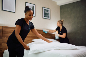 Smiling black maid changing bed linen while cleaning hotel room with female co-worker.