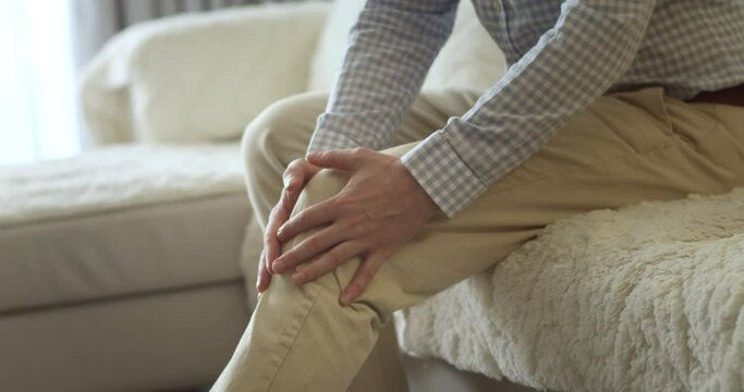 Senior european man suffers from painful severe knee joint pain, could not walk, illness, cramps, rheumatism.Close up.Male hands massaging knee leg muscles while sitting on sofa in living room at home
