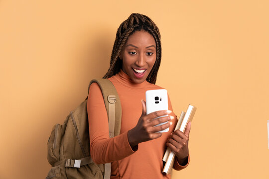 joyful college student watching video and photos on mobile smartphone in beige studio background. dedication, effort education concept.