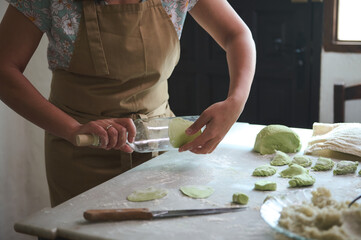 Close-up housewife using glass wine bottle as rolling pin, rolling out green spinach dough for making dumplings, standing at kitchen table sprinkled with flour in the rural house kitchen