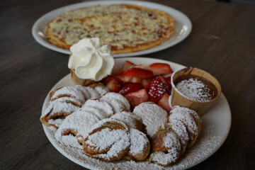 a plate of poffertjes, a traditional Dutch pancake, covered in sugar powder with a side of strawberry fruit and chocolate sauce. A savory pancake with a cheese, bacon, and egg in the background