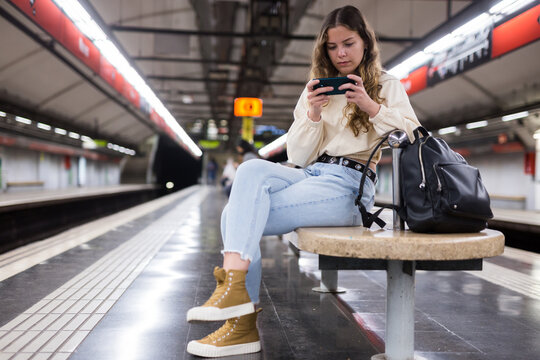 Portrait of a focused girl sitting on a subway platform bench, texing with friends on a mobile phone