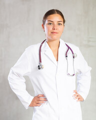 Friendly young female professional doctor in white coat posing against gray studio background, looking at camera with smile