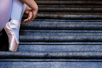 Closeup of ballerina wearing pointes shoes holding her hands on her ankles while sitting on the gray stone steps. Dance resting on the stair. Dancer´s feet with pink ballet slippers on. Copy space - obrazy, fototapety, plakaty