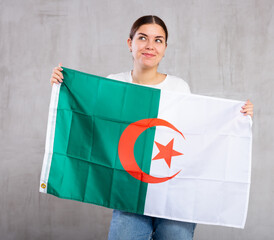 Joyous young woman with Algeria flag in hands posing happily against light unicoloured background