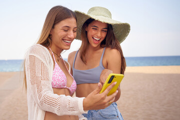Two beautiful best friends looking at cell phone having fun on beach. Young happy women in swimwear...