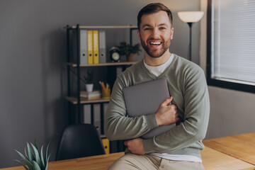 Modern happy bearded office male worker of IT company smiling and holding laptop in office