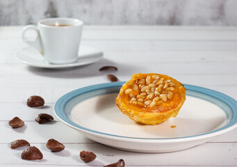 Close-up of a cream and pine nut tartlet with chocolate flakes and a cup of coffee.