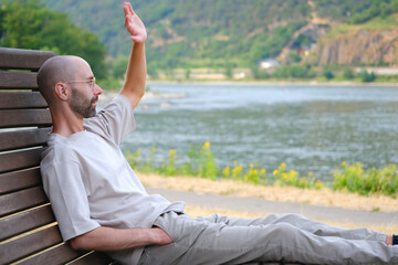 young man 30 years old sites comfortably on wooden park bench on banks of river, lake, resting on...