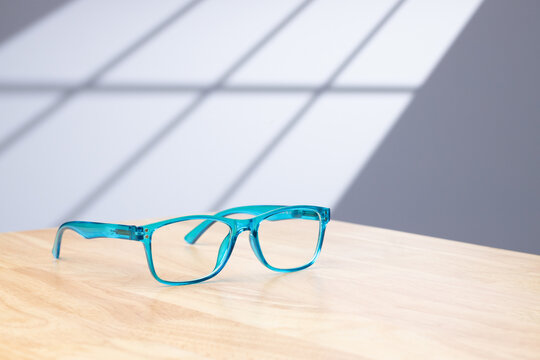 a pair of blue reading glasses on a light wood desk in a home office with sun light casting a window shadow on the background.
