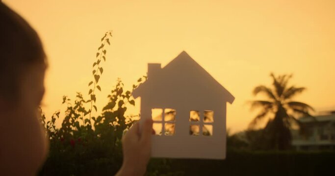 Paper house, sun shines thruth of window paper house.Child hand holding paper house on background sunset. Symbol home, family comfort, happy. Dream buy house. Real estate insurance.