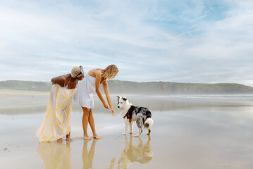 multiracial pair of female friends interacting with their border collie dog while taking a walk along the seashore at the beach.