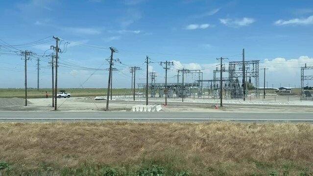 The camera pans over an electrical substation in the San Joaquin Valley region of California. Electrical poles, electrical wires, and other infrastructure dominate the landscape.
