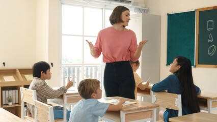 Caucasian teacher teaching and explaining theory to multicultural student by using body gesture while standing at blackboard in elementary school. Skilled teacher asking children question. Pedagogy.