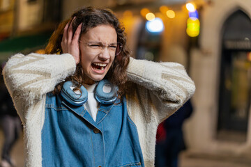 Displeased angry young woman frowns face feels disgust suffering from loud sound covers ears with hands palms refusing to listen gossip bad information don't hear. Teen girl stand on street at night.