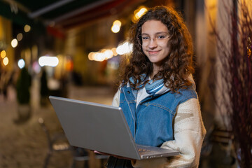 Happy teenager girl working online distant job with laptop in city street, browsing website chatting outdoors. Caucasian young woman tourist looking at camera thinking typing in town at night