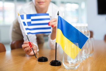 Female hands arranging flags of Greece and Ukraine on table before international summit meeting,...