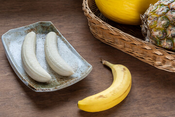 Exotic fruits on the wooden surface. A ceramic plate with two peeled bananas, a wicker basket with...