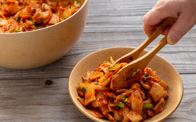 A hand putting kimchi on a plate with wooden tongs. A traditional Korean side dish of salted and fermented vegetables with garlic, ginger, and gochugaru. 