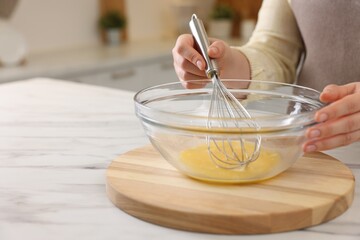 Woman whisking eggs in bowl at light marble table indoors, closeup. Space for text
