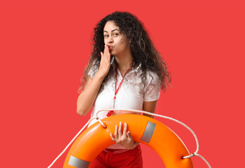 Beautiful young African-American female lifeguard with ring buoy blowing kiss on red background