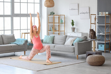 Young pregnant woman doing yoga with cute cat scratching post at home