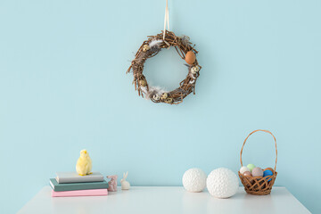 Beautiful Easter wreath on blue wall near table with basket of eggs in room