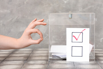 Voting woman showing ok gesture near ballot box on table against grey background