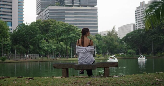 Back view of woman sitting on bench in Bangkok park water lake and skyscrapers behind. Bangkok park urban green space Bangkok park reflecting Thailand's natural charm Haven of tranquility.