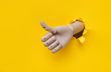 A wooden hand protrudes from a torn hole in yellow paper and shows a big thumbs up like gesture....