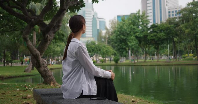Woman sits thoughtfully on bench in city park near lake side view close-up skyscrapers in background. City park fusion of urban and natural beauty City park tranquil space for reflection.