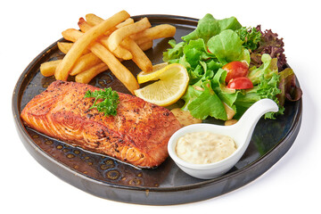 Large salmon steak served with fried french fries, fresh salad and tomatoes, creamy dipping sauce served on black plate isolated on white background.