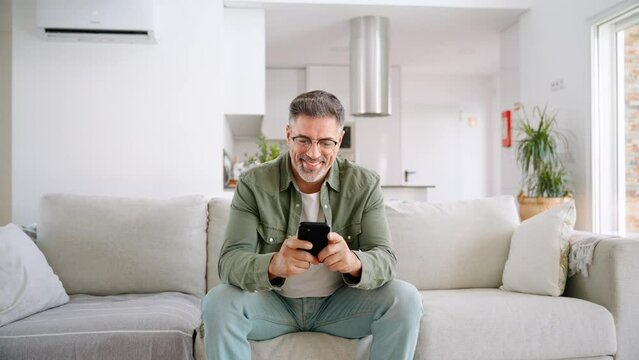 Relaxed happy older mature middle aged man holding mobile phone using cellphone sitting at home on sofa in modern living room, chatting online, texting messages, reading news, shopping in apps.
