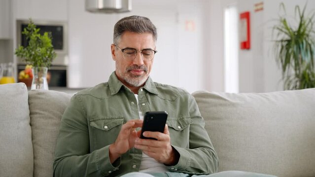 Happy relaxed mature man using cellphone relaxing on couch at home. Middle aged old user holding smartphone browsing internet, texting messages on mobile cell phone technology sitting on sofa.