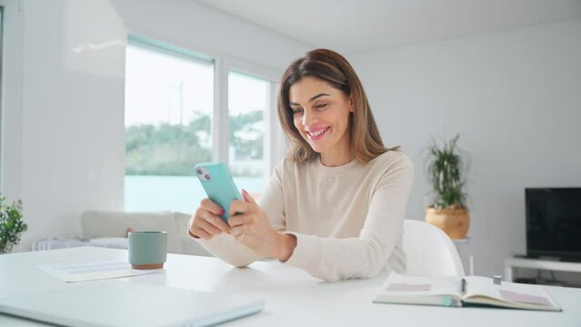 Happy mature woman using mobile phone sitting at home table. Smiling older middle aged lady holding smartphone scrolling buying online doing shopping in smartphone app in living room. Copy space.
