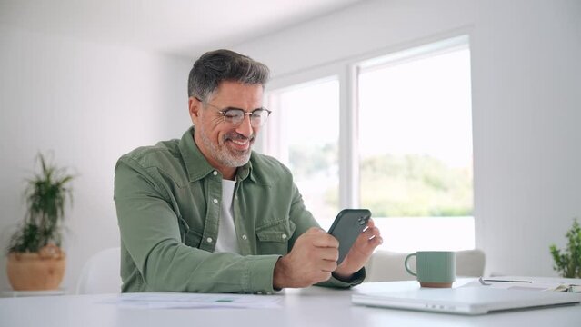 Happy senior older man using smartphone at home. Smiling middle aged mature user looking at mobile cell phone technology applications making banking payments sitting at table in living room.