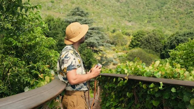 a male traveler in a safari hat takes a photo on a suspension bridge. traveler with backpacks relaxing in greens jungle and enjoying view in waterfall. tourist traveling along rocky forest trek.