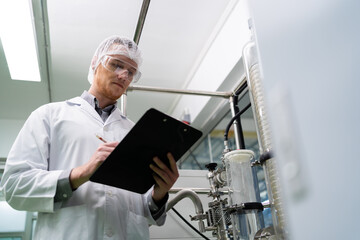 A scientist wearing a lab coat and goggles is writing on a clipboard. The clipboard is black and the scientist is looking at it intently