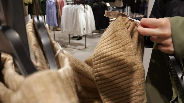 A girl reads a sweater label in a clothing store. The girl chooses a warm sweater.