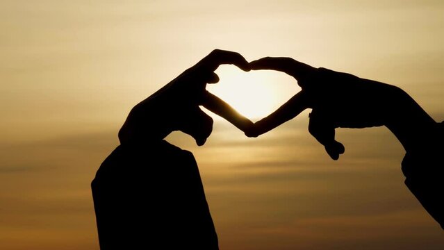 Women's hands depict the shape of a heart against the background of a heavenly sunset. The fingers are arranged in the shape of a heart. A beautiful romantic scene.